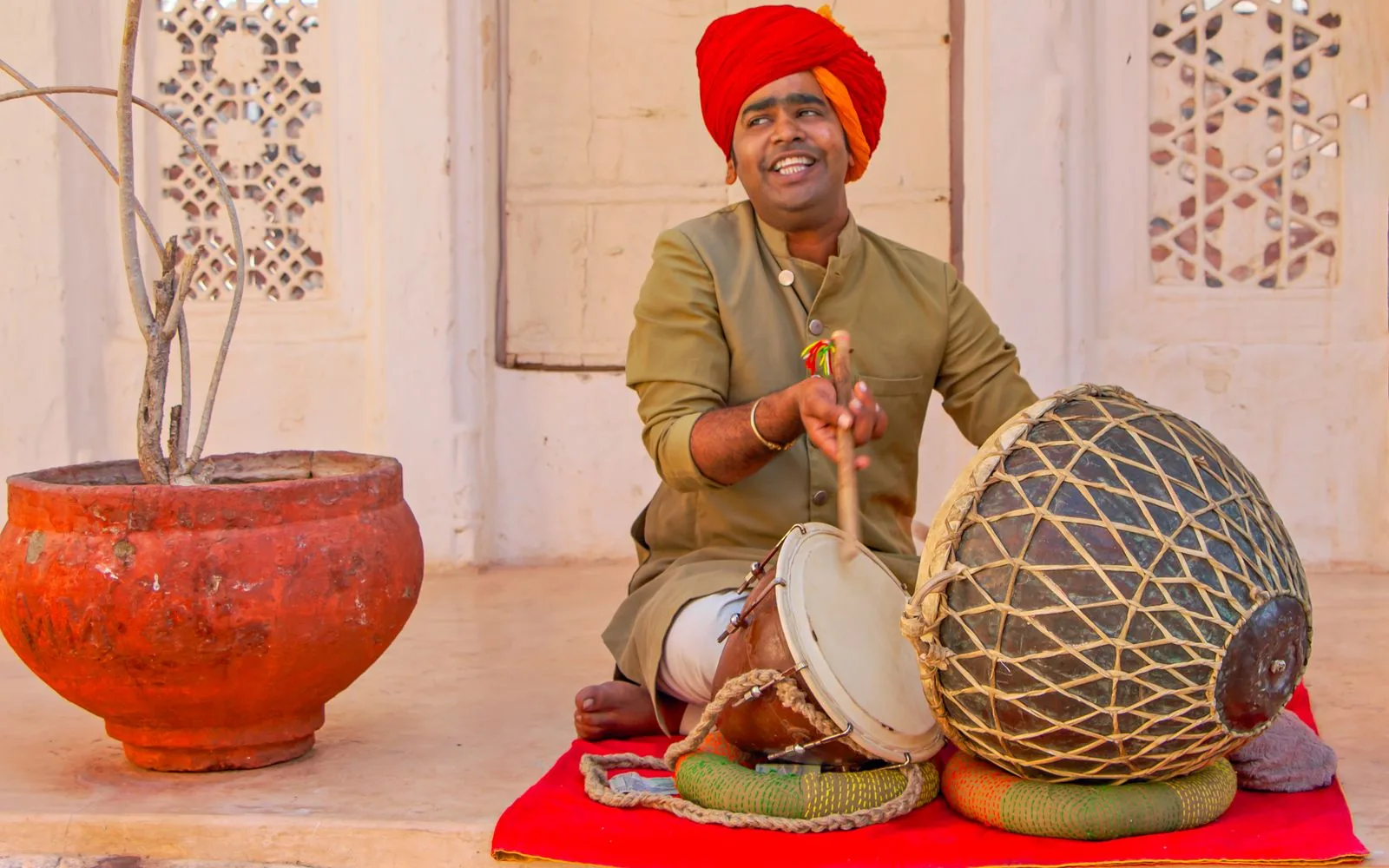 A smiling Rajasthani percussionist playing traditional drums on a red carpet before carved jharokha doors