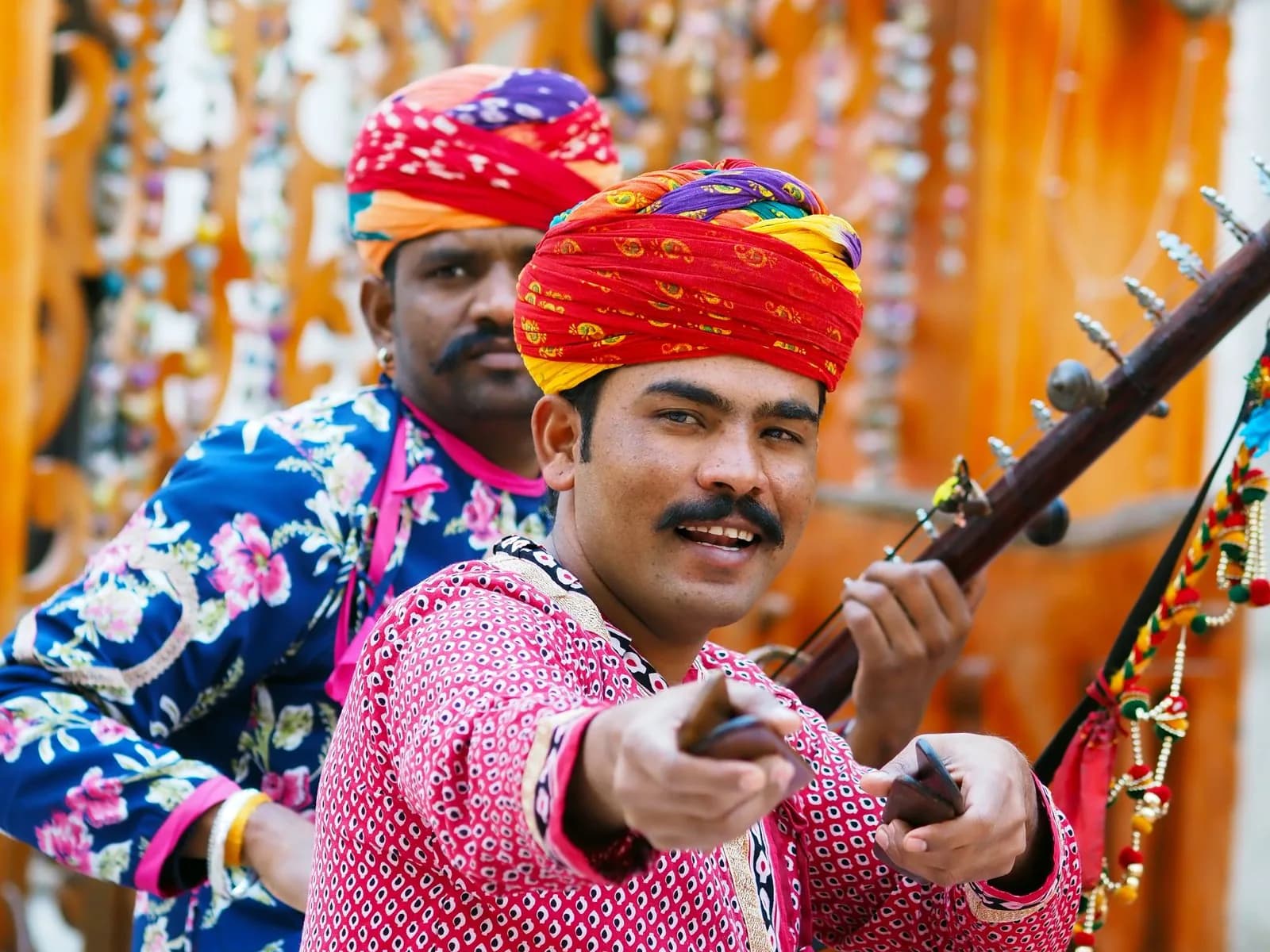 Two Rajasthani folk musicians in vivid turbans singing with tanpura and khartal against warm carved wood